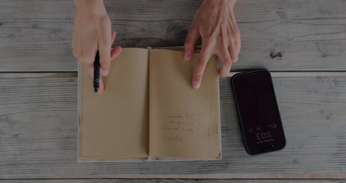 Overhead View Of Hand Opening Notebook Beside Smartphone On Wooden Table, Capturing Start Of Journaling Or Planning Session