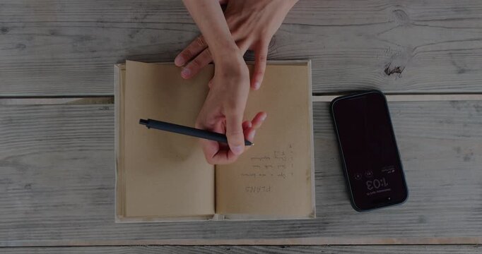 Overhead View Of Person Writing Plan In Open Notebook Beside Smartphone On Wooden Table, Showing Organization And Daily Productivity