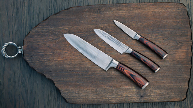Three kitchen knives placed on a wooden cutting board with a ring on the side in a kitchen setting during the day