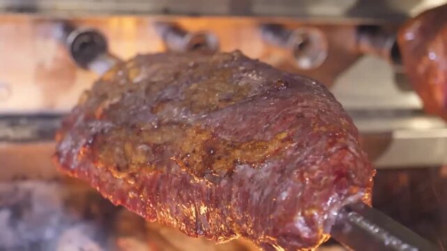 Close-up footage of a large, juicy cut of picanha beef cooking on a metal skewer over hot charcoal flames in a traditional Brazilian steakhouse, or churrascaria. The meat is seasoned and sizzling, rea