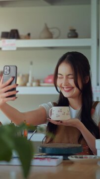 Happy asian female artist taking selfie with handmade pottery design in creative studio &ndash; smiling sculptor sharing artwork on social media for online selling, digital portfolio and creative expression