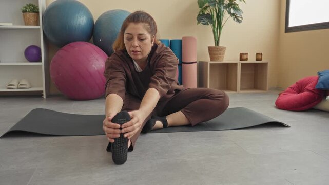 Woman seated on a yoga mat reaches forward and grasps her foot with both hands, stretching hamstring and ankle in a studio; serenity mindfulness.