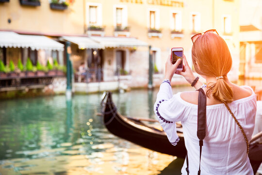 Rear view of a young Woman standing by a canal in summer taking a photo with her mobile phone, Venice, Veneto, Italy