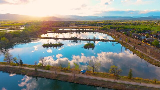 Aerial drone flies forward over glassy Colorado ponds reflecting clouds at golden sunset with Rocky Mountain backdrop neighborhood and walking trail