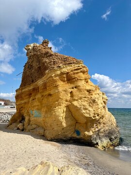 A scenic view of a massive, textured yellow sandstone cliff on the edge of a sandy beach against a blue sky with white clouds.