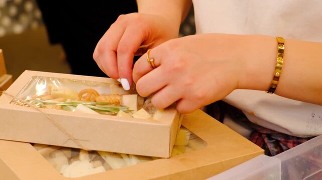 Female seller hands in wine shop tie ribbon on package with assortment for wine snack from different cheese types and crisp unsweet cookies and crispbread. Wine snack. Wine festival.
