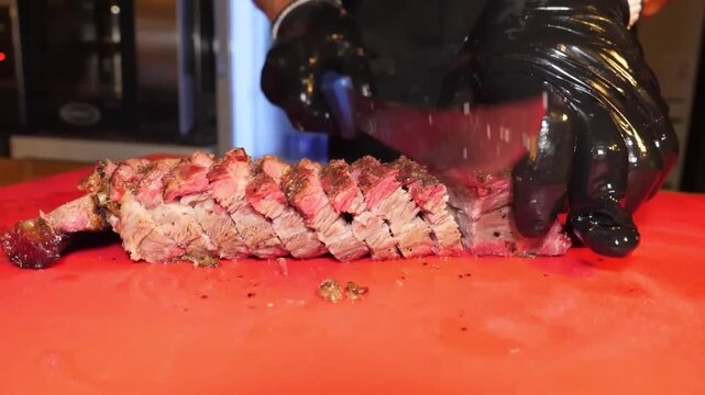 Close-up of a chef wearing black gloves using a carving knife to slice a perfectly cooked, juicy sirloin rump roast. The meat is resting on a red cutting board in a professional kitchen or restaurant 