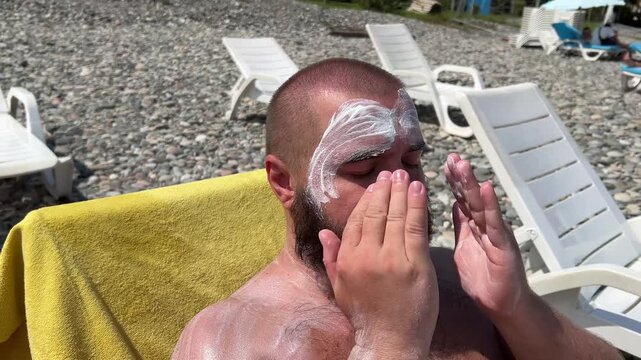 Travel video. 60 fps. Young adult tanned bald white fat naked man with beard applies sunscreen cream, moisturizer, milk balm to face on sun lounger with yellow towel on ocean coast, sea beach. Tourism