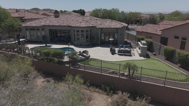 A 4k aerial view of a desert landscaped home in Arizona featuring a travertine tiled pool deck with a fireplace and outdoor kitchen.