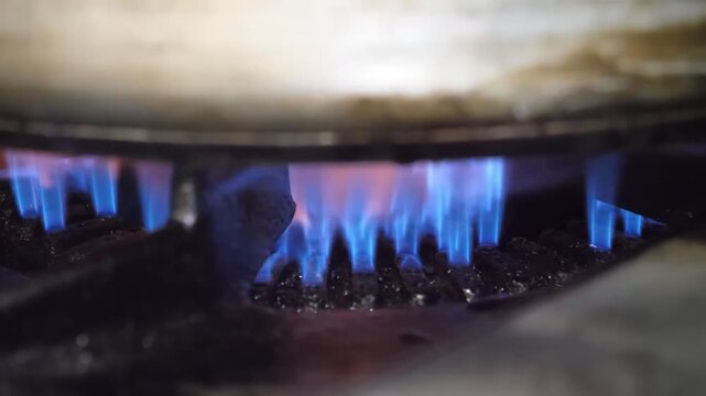 A detailed, eye-level shot of a gas stove burner with blue and orange flames heating the bottom of a large pot or pan. The scene captures the heat, energy, and process of cooking in a kitchen environm
