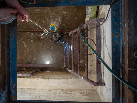 Hands lowering electric water drainage pump with rope and float switch into dark flooded concrete basement pit through narrow metal hatch featuring rusty ladder and hose pipes.