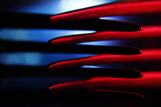 Tenedores met&aacute;licos dispuestos en superposici&oacute;n, iluminados con luces rojas y azul que generan un efecto abstracto. Las l&iacute;neas repetitivas de los dientes crean un patr&oacute;n visual con sombras.   