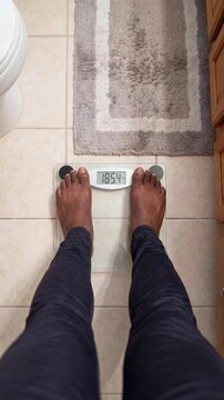 Top view of person's feet stepping onto digital scale in home bathroom with the screen indicating a weight measure of 185.4 lbs