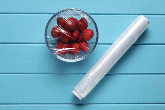 Bowl with fresh strawberries wrapped in cling film and roll of plastic wrap on blue wooden table, flat lay