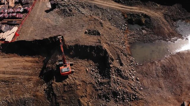 Aerial shoreline construction site with excavator shoveling ground. Machinery is operating at water edge with puddles. Active trenching and runoff control. Work equipment is engaged in earth job