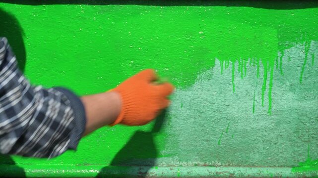 Individual in plaid shirt and orange gloves paints a green surface, showcasing the gradual application and blending of paint in a well-lit outdoor setting