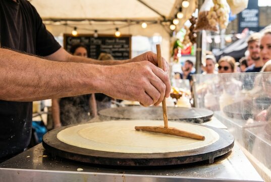 Street food vendor hands using a traditional wooden t shaped spreader to smooth crepe batter on a hot circular griddle