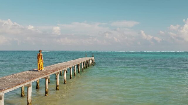 Woman in yellow walks along a wooden pier over turquoise ocean in Mexico