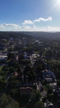 Vista a&eacute;rea de cidade cercada por vegeta&ccedil;&atilde;o, com bairros, casas e colinas no horizonte sob luz intensa do sol, criando paisagem urbana ampla em atmosfera de fim de tarde.
