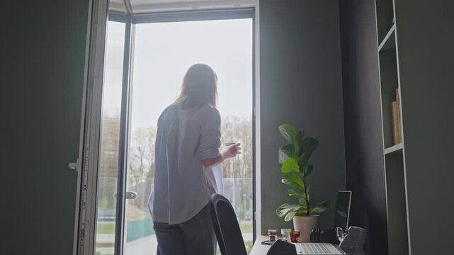 Woman holding coffee cup and opening balcony door while looking outside. Concept of morning routine and break