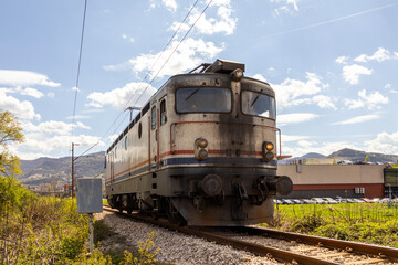 Old electric locomotive moving along railway tracks in rural landscape, front view capturing motion and power, with mountains and buildings in background under bright blue sky. © Daanree