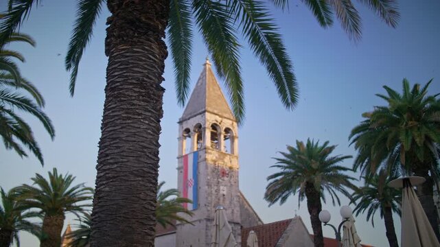 morning establishing shot of the 14th-century bell tower of the Church and Monastery of St. Dominic in Trogir, Croatia. The stone tower features a waving Croatian flag, framed by lush palm trees again