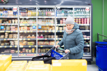 Obraz premium Mature woman in warm jacket and knit hat pushing shopping cart inside brightly lit supermarket aisle with frozen food section.
