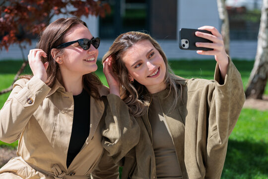Two young women take a selfie together while sitting on a sunny day outside in a green park area
