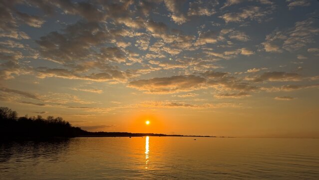 Golden sunset sky with scattered clouds over a calm sea. Bright sun reflecting on the water surface with dark forest silhouettes on the coast.