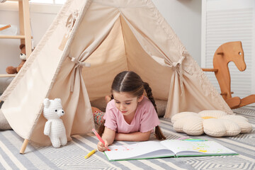 Little girl drawing in sketchbook near toy wigwam at home © New Africa