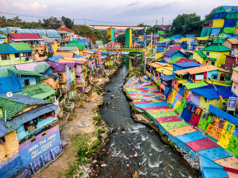 Aerial view of the vibrant Kampung Warna Warni Jodipan with people walking across the bridge and colorful houses along the Brantas River in Kecamatan Blimbing, Jawa Timur, Indonesia.