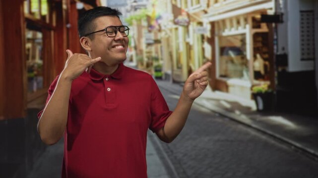 Hispanic man makes call me hand gesture and points finger on street wearing glasses and red polo; playful greeting.