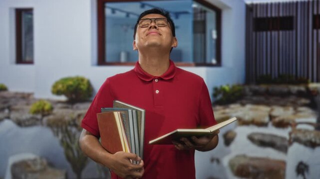 Young hispanic man wearing glasses holding multiple books and reading from an open book at a building entrance outdoors; study concentration.