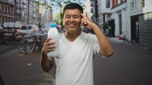 Man holding a plastic bottle points finger to temple on street, smiling with eyes closed; thinking confidence.