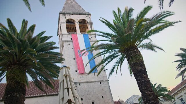 morning establishing shot of the 14th-century bell tower of the Church and Monastery of St. Dominic in Trogir, Croatia. The stone tower features a Croatian flag, framed by lush palm trees