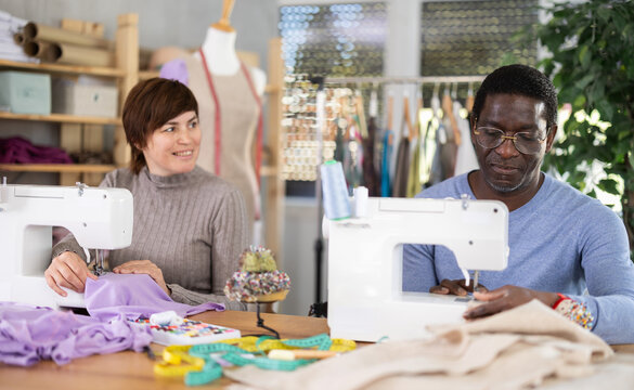 Positive male and female dressmakers stitching garments at machines, working together in small cozy sewing workshop filled with fabrics and tools
