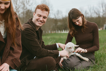 A group of young adults enjoy a sunny day in a rural field, gently stroking a small goat kid. A...