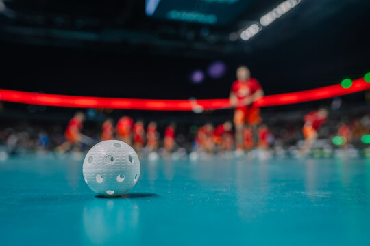 Close-up of a white floorball on a turquoise court with players in red uniforms blurred in the background