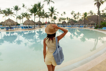 Fototapeta premium Woman standing by the pool at resort, looking at the water with palm trees and chairs on background, back view