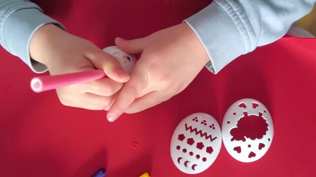 A child colors an Easter egg with a pink marker using a plastic stencil.