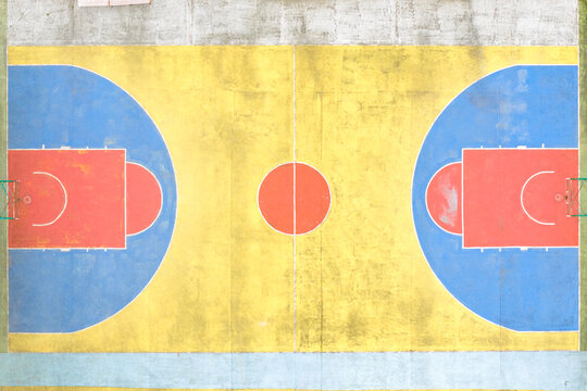 Aerial view of a colorful outdoor basketball court with yellow, blue, and red painted surfaces and white markings in San Giovanni Valdarno, Tuscany, Italy.