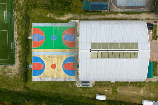 Aerial view of colorful outdoor basketball courts and a large arched gymnasium roof surrounded by green fields in San Giovanni Valdarno, Tuscany, Italy.