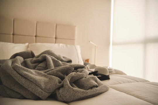 Cozy unmade bed with a gray blanket, white pillows, ambient morning light, and headset