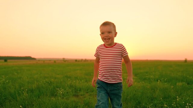 Kid laughs runs across green grass, sunset. Small child plays on lawn. Child active games. Happy boy laughs while play on lawn. Small boy runs green grass with arms raised. Family picnic, kid nature