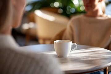 Fototapeta premium White coffee cup on table with two women in cozy cafe setting