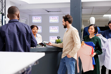 Obraz premium Man standing in line making contactless smartphone payment, paying for purchases with electronic credit card while shopping on Black Friday. Diverse people holding clothes waiting at cash regiter desk