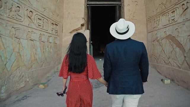 american young man traveler wearing white hat with his wife with red dress exploring ancient Egyptian abu simble temple with background of ramsses ii statues. Egypt.
