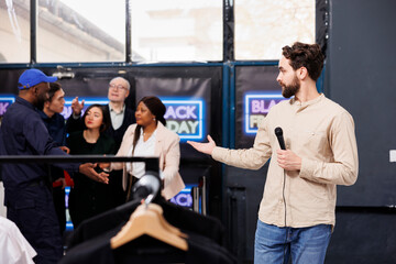 Obraz premium Man TV reporter standing at shopping mall entrance presenting news about Black Friday. Clothing store security guard opening door for eager people shoppers waiting for discounts and seasonal sales