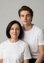 Obraz premium Happy woman and her son smiling at camera. Mother and young man standing together against grey background. Portrait of Caucasian family members dressed in simple white t-shirt clothes.