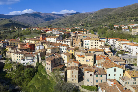 Aerial view of historic stone buildings and terracotta rooftops nestled against the lush green hills of the Pratomagno mountains under a clear blue sky in Loro Ciuffenna, Tuscany, Italy.